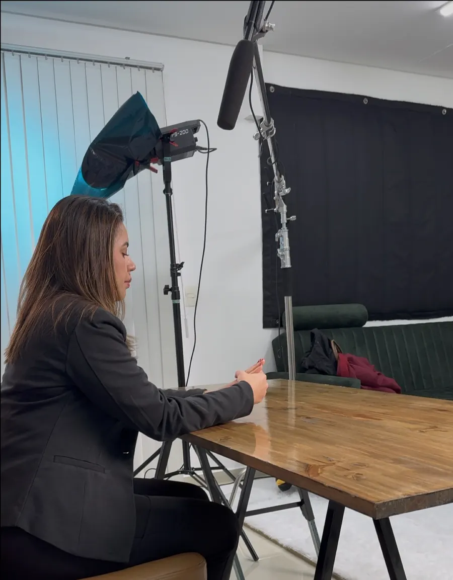 Gravação na mesa de madeira do estúdio, com microfone boom suspenso, iluminação Amaran e sofá verde ao fundo