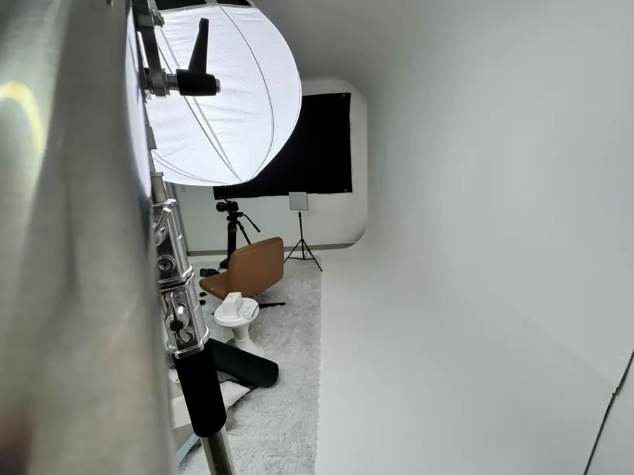 Sala do estúdio montada para gravar, com lanterna difusora, câmera em tripé e poltrona sobre o tapete branco