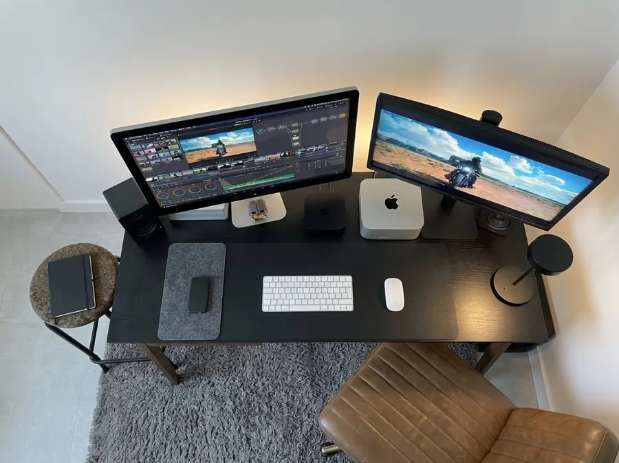 Ilha de edição vista de cima, com dois monitores, Mac, teclado e mousepad sobre a bancada preta